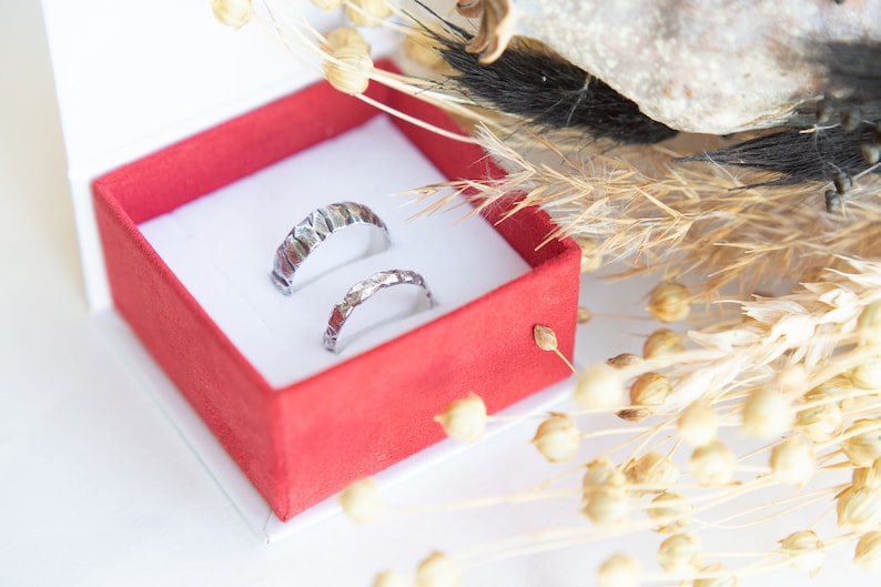 May include: Two silver wedding bands with a textured finish, displayed in a red velvet ring box.