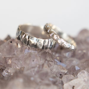 May include: Two silver wedding bands with a textured hammered finish, resting on a bed of amethyst crystals.