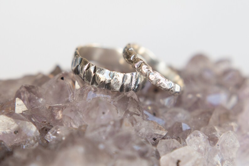May include: Two silver wedding bands with a textured hammered finish, resting on a bed of amethyst crystals.