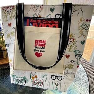 May include: White canvas tote bag with black straps and a large pocket. The bag is printed with a pattern of dogs wearing glasses. The pocket has the text "Beware of Dogs They Will Steal Your" with a red heart.