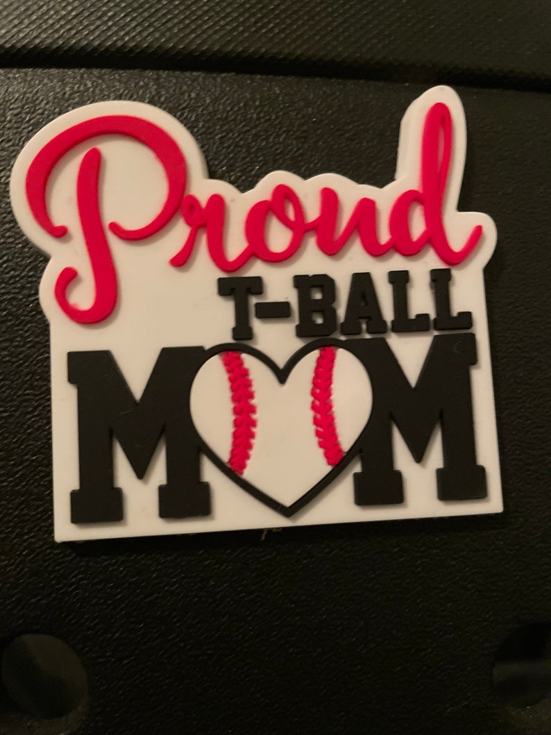 May include: A white and black rectangular badge with the words "Proud T-Ball Mom" in red and black lettering. The word "Proud" is in a cursive font. The letter "O" in "MOM" is a baseball-shaped heart.