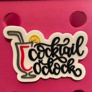 May include: A white, black, red, and yellow "Cocktail O'Clock" charm. The charm features a cocktail glass with a straw and a lemon slice. The charm is set against a pink background.