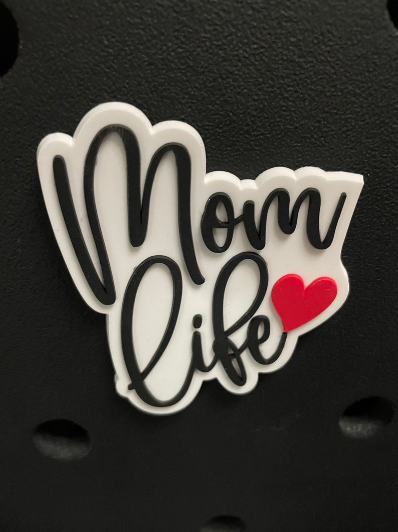May include: A white and black "Mom Life" charm with a red heart accent. The charm is in a cloud-like shape and is set against a black background. The text is in a cursive font.
