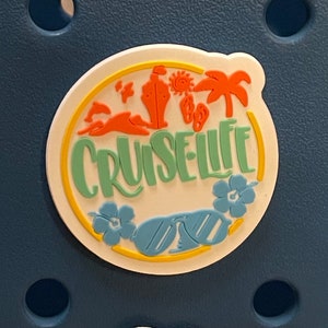 Pu&ograve; includere: Un ciondolo circolare bianco con la scritta "CRUISE LIFE" in verde, circondato da elementi grafici a tema spiaggia arancioni e gialli. Include occhiali da sole, fiori, una nave da crociera e palme. Il ciondolo &egrave; posto su uno sfondo blu scuro.