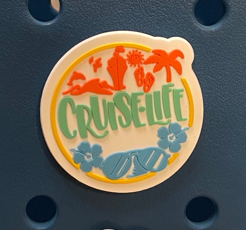 Pu&ograve; includere: Un ciondolo circolare bianco con la scritta "CRUISE LIFE" in verde, circondato da elementi grafici a tema spiaggia arancioni e gialli. Include occhiali da sole, fiori, una nave da crociera e palme. Il ciondolo &egrave; posto su uno sfondo blu scuro.