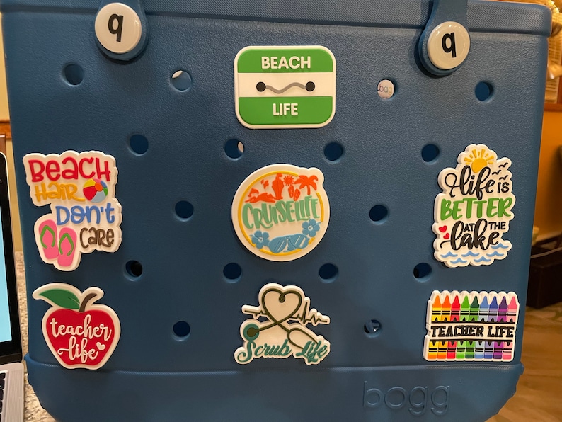 Pu&ograve; includere: Una borsa tote blu scuro adornata con vari adesivi colorati. Gli adesivi presentano frasi come "Beach Life", "Beach Hair Don't Care" e "Teacher Life". Altri adesivi raffigurano una scena di crociera, uno stetoscopio e un lago.