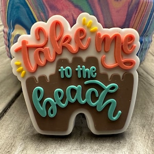 Puede incluir: Un adorno de zapato colorido con la frase "take me to the beach" en coral, turquesa y marrón. El adorno tiene forma de castillo de arena y tiene detalles amarillos. El fondo es una mezcla borrosa de colores.