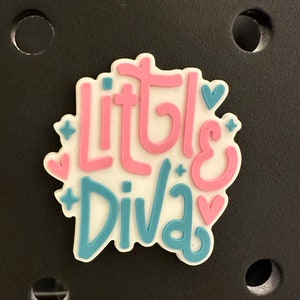 May include: A white, pink, and blue charm with the words "Little Diva" in a playful font. The charm features pink hearts and blue star accents. The charm is set against a black background.