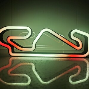 Könnte beinhalten: Ein Neonlicht in Form einer Rennstrecke mit schwarzer Umrandung und weißer und roter Innenbeleuchtung. Das Licht spiegelt sich auf einer glänzenden Oberfläche und erzeugt ein Spiegelbild. Das Farbschema ist grün und rot.