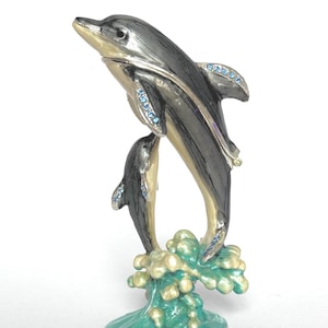 May include: A decorative figurine of two dolphins leaping from a wave. The dolphins are silver and black with blue gemstone accents. The wave is green and gold. The figurine is on a white background.