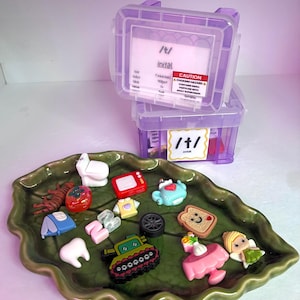 May include: A green leaf-shaped ceramic tray holding various small plastic figurines including a white toilet, red television, blue tent, pink tooth, green tank, black wheel, smiling toast, and fairy, with two purple storage bins labelled /t/.