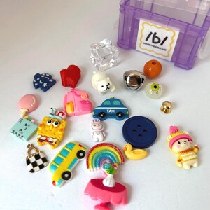 May include: A collection of colorful craft embellishments, including a yellow SpongeBob SquarePants, a blue taxi car, a rainbow, a pink backpack, a blue button, and a clear cube. Other items include a white dog, a bell, and a small bear.