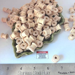 May include: A large collection of small, light-colored wooden spools piled in a green leaf-shaped dish. A stainless steel ruler is positioned at the bottom, showing measurements in inches. A small rectangular sign reads CAUTION CHOKING HAZARD.
