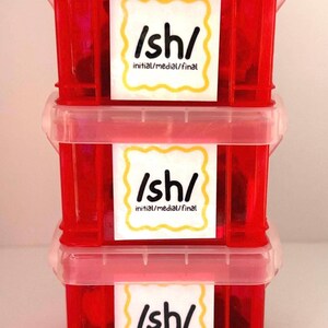 Può includere: Tre contenitori impilati di plastica rossa con coperchi trasparenti. Ogni contenitore ha un'etichetta bianca con il testo "/sh/ initial/medial/final" in nero, bordata di giallo. I contenitori sono probabilmente per scopi educativi.