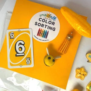 May include: A yellow color sorting kit featuring a card with the number six, a yellow tassel, a small yellow bee figure, and a circular label reading smalltalk COLOR SORTING THEMED MINI OBJECTS. The set includes various yellow miniature items.