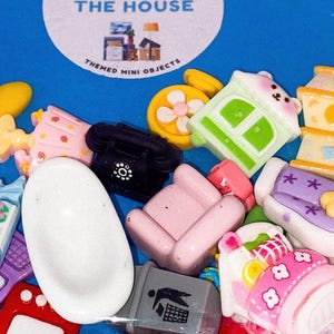 May include: A collection of themed mini objects, including a white bathtub, a black rotary phone, a pink armchair, and a green house-shaped charm. The objects are scattered on a blue surface, with a circular label that reads "Around the House."