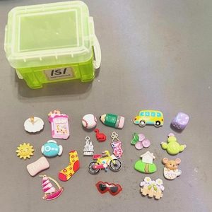 May include: A collection of colorful miniature charms and a green plastic storage container. Charms include a baseball, pencil, school bus, dice, and a bicycle. Other charms include a sailboat, sun, and a pair of sunglasses.
