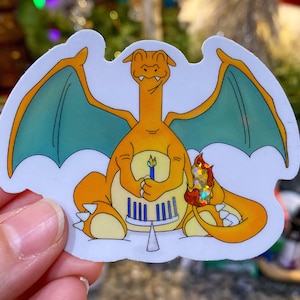 May include: A sticker featuring a cartoon Charizard character with orange scales and teal wings. The character is holding a birthday cake with blue candles and a lit candle in its other hand. The sticker has a white border.