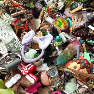 May include: A dense collection of assorted craft supplies and trinkets including a lavender gingham hair tie, a Rocket Grape Soda bottle cap, a silver thimble, a pink resin ring, a brown gingerbread-shaped charm, and a metal butterfly charm.