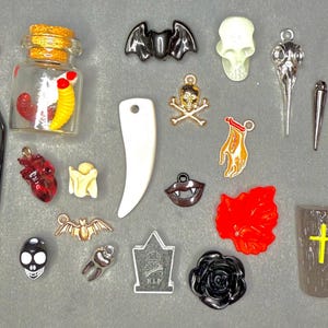 May include: A collection of Halloween-themed charms and pendants in various shapes and colors. Includes a black coffin, a skull, a bat, a tooth, a red rose, a jar with worms, and a knife with floral details.
