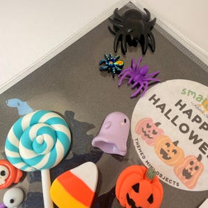 May include: A collection of miniature Halloween-themed charms including a black spider, a metallic blue spider, a purple spider, a light purple ghost, a bright orange pumpkin, a candy corn piece, and a blue and white swirl lollipop on a stick.