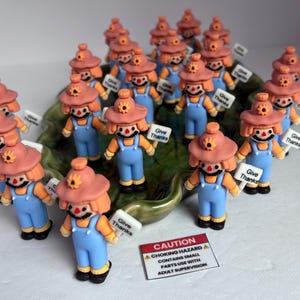 May include: A collection of small scarecrow figurines, each wearing a peach-colored hat with a yellow flower, orange hair, and light blue overalls. Many hold a white sign that reads Give Thanks. A rectangular caution label is placed in the front.