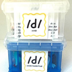 Op de afbeelding: Twee doorzichtige plastic opbergdozen op elkaar gestapeld. De bovenste doos is transparant, de onderste blauw. Elke doos heeft een wit label met "/d/" en "initial" of "initial/medial/final".