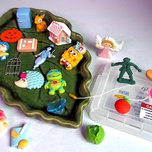 May include: A collection of miniature plastic charms and figurines including a green leaf-shaped tray, a blue refrigerator, a pumpkin, a gingerbread house, a turtle, a sponge character, an angel, a green soldier, and a bandage-shaped item.