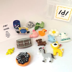 May include: A collection of miniature toys and objects, including a blue die, a toy drum, a donut, a duck, and a deer. The image also includes a card with the text "/d/ initial" and a list of words.