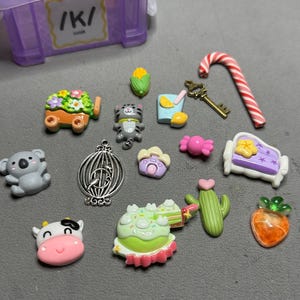 May include: A collection of colorful miniature charms and embellishments, including a candy cane, a key, a cat, a koala, a cow, a cactus, a carrot, a cake, and a birdcage. A purple storage box is in the background.