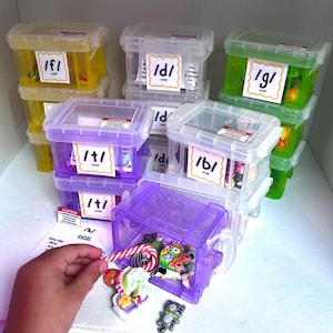 May include: A collection of small, stackable, translucent plastic storage bins in yellow, clear, purple, and green. Each bin features a white label with phonetic symbols like /f/, /d/, /g/, /t/, and /b/. One purple bin is open, revealing miniature toy figurines.