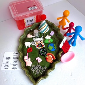 May include: A green leaf-shaped ceramic tray containing various small objects including a thimble, a party hat, a compass, a turkey figurine, a tooth, a moth charm, and a Thor character. A red plastic storage box and three colorful figures are nearby.
