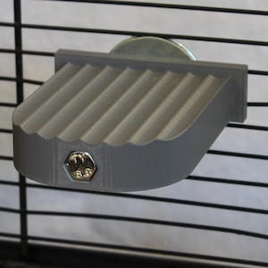 May include: A gray, ridged pet cage platform attached to black wire mesh. The platform has a curved edge and is secured with a silver bolt. The platform is designed to provide a resting spot for small animals.
