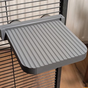 Puede incluir: Una estantería gris con ranuras para jaula de mascotas, fijada a una jaula de alambre. La estantería es rectangular con esquinas redondeadas y está hecha de un material sólido y duradero. Está diseñada para proporcionar un área de descanso o juego para mascotas pequeñas.