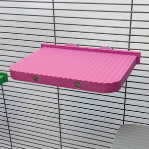 Può includere: Una mensola rettangolare rosa brillante con una superficie testurizzata e ondulata, dotata di due bulloni metallici visibili sul bordo anteriore. La mensola è fissata a una griglia metallica scura tramite due ventose circolari sul retro.