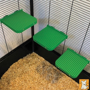 May include: Three square green platforms with a ribbed surface texture, designed to be mounted to the interior wire walls of a pet enclosure. Each platform features small circular attachment points along the back edge for secure installation.