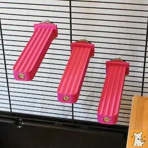 May include: Three bright pink, rectangular, ribbed platforms attached to a black wire cage wall. Each platform features a flat mounting end with a visible metallic bolt head at the front, extending horizontally to provide a stepped perch surface.