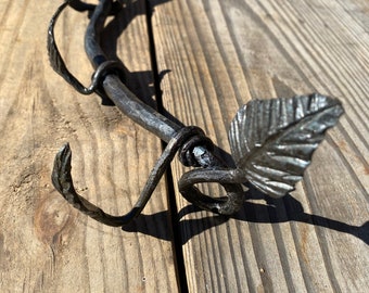 May include: A black metal coat rack with three hooks and leaf-shaped accents. The rack is curved and has a rustic, handmade look.