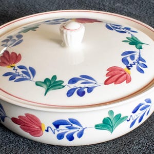 Op de afbeelding: Een witte keramische ovenschaal met deksel, met een bloemmotief in rood, blauw en groen. De schaal heeft handvatten en een knop op het deksel. De rand is geaccentueerd met een rode lijn. Een decoratief keukenartikel.