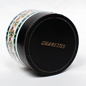 May include: A black cylindrical cigarette case with the word "CIGARETTES" in gold lettering. The case has a decorative band with a floral pattern in green, white, and red. The case is made of leather.