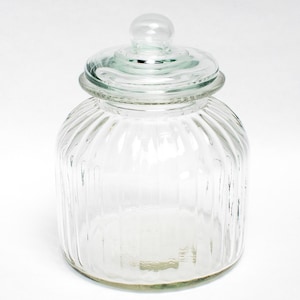 Op de afbeelding: Een doorzichtige glazen voorraadpot met verticale ribbels langs het lichaam. De pot is voorzien van een bijpassend afneembaar glazen deksel met een kleine ronde knop als handvat aan de bovenkant. De houder heeft een brede ronde opening.