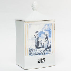 Op de afbeelding: Witte keramische pot met deksel en decoratieve knop. De pot heeft een blauw-witte illustratie van mensen in een landelijke omgeving. Het woord "SUIRER" is in het zwart op de voorkant van de pot gedrukt.