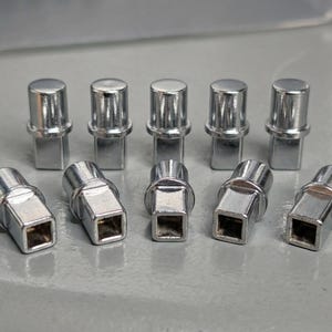 May include: Ten shiny, chrome-coloured lug nuts are displayed. Five have a cylindrical top and a square base, standing upright. The other five are angled, with a square base and a hexagonal top. Automotive hardware.