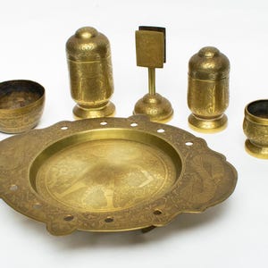 Op de afbeelding: Een verzameling antieke messing items, waaronder een grote decoratieve schaal, twee potten met deksel, een kleine kom, een kleine beker en een standaard met een rechthoekige houder. De items hebben ingewikkelde geëtste ontwerpen.
