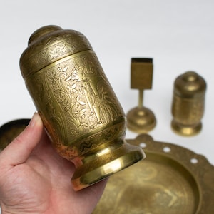 Op de afbeelding: Een verzameling antieke messing items, waaronder een decoratieve container met een deksel, een kleine lucifershouder, een zoutstrooier en een decoratieve schaal. De container wordt in een hand gehouden en heeft een gegraveerd ontwerp.