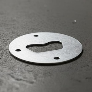 Op de afbeelding: Een close-up van een zilveren metalen ring met een sleutelgatvormige uitsparing en drie kleine gaten. Andere metalen componenten zijn wazig op de achtergrond, alles op een getextureerd grijs oppervlak.