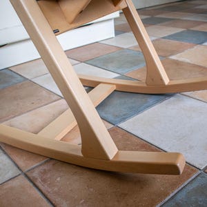 May include: Close-up of the base of a wooden high chair. The chair has a natural wood finish and is designed with curved legs and a rocking base. The chair is positioned on a tiled floor with various colors.