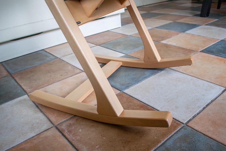 May include: Close-up of the base of a wooden high chair. The chair has a natural wood finish and is designed with curved legs and a rocking base. The chair is positioned on a tiled floor with various colors.