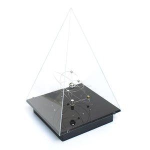 Op de afbeelding: Een kinetisch sculptuur met een centraal metalen orbitaalmodel ingesloten in een doorzichtige acryl piramide. De basis is een vierkant zwart platform met kleine zilveren stippen, dat een reeks in elkaar grijpende draadringen ondersteunt.