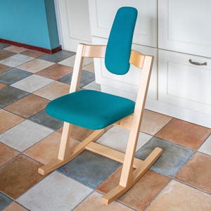 May include: A teal ergonomic chair with a wooden frame. The chair has a curved backrest and seat, and a rocking base. The chair is set against a white cabinet and a tiled floor.